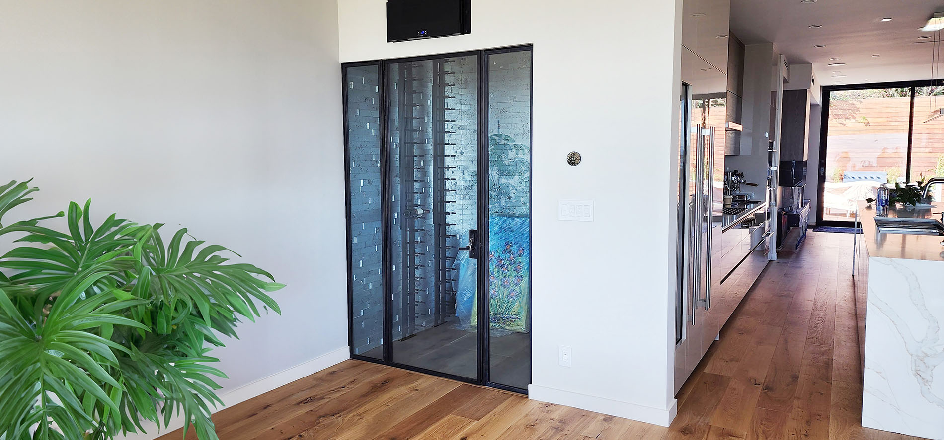 This image shows a stylish wine cellar just off the kitchen, enclosed by sleek glass doors with black frames. To the left, a green plant adds a natural touch, balancing the modern design with organic warmth. The glass enclosure offers a clear view of the wine storage, blending functionality with elegant aesthetics.